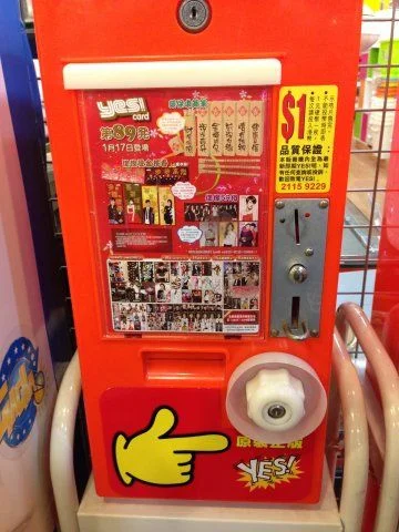 An orange YES! Card vending machine in a Hong Kong shop, with a coin slot, a white turning knob, and a big yellow pointing hand.